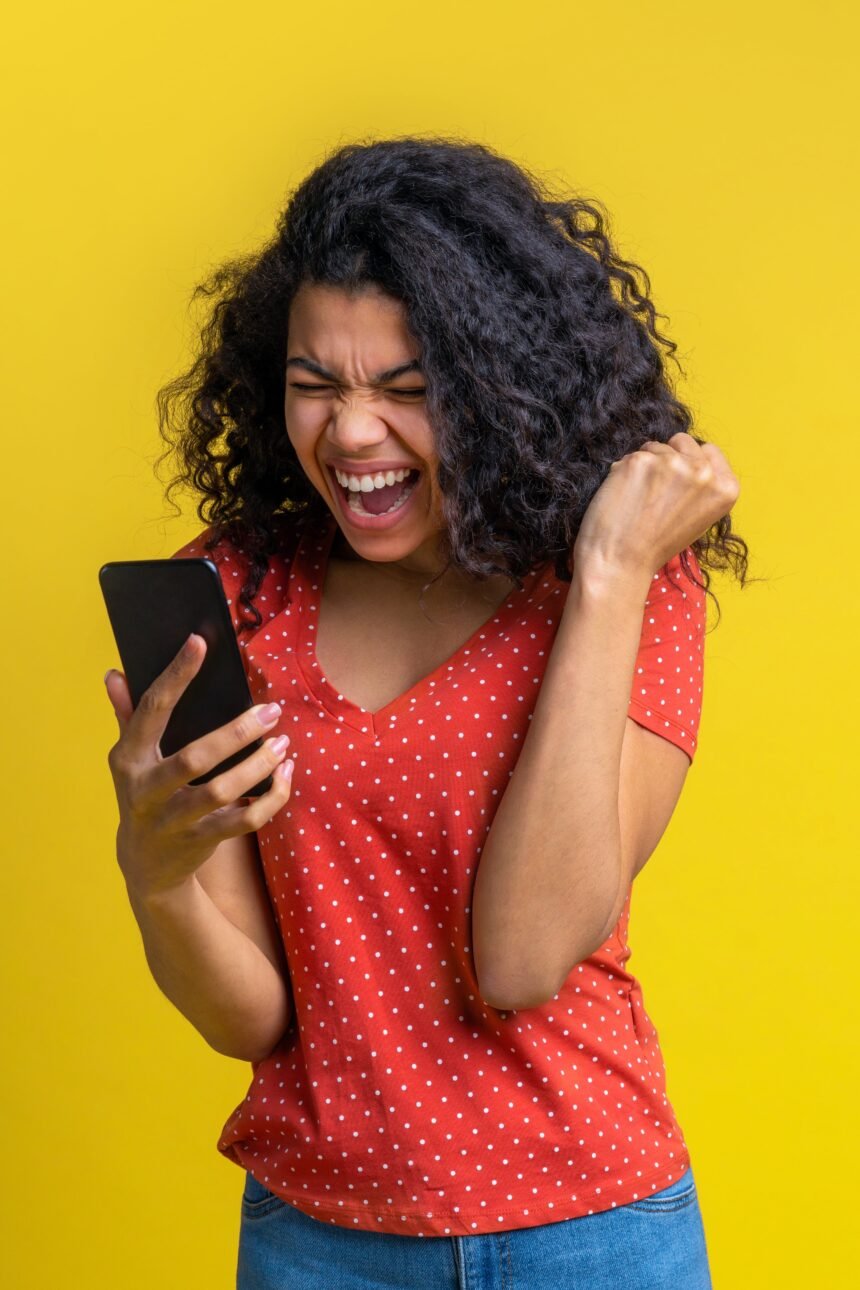 Beautiful dark skinned girl holding mobile phone and making winner's gesture clenching her fist, screaming yes celebrating success, expressing joy about victory in online lottery.