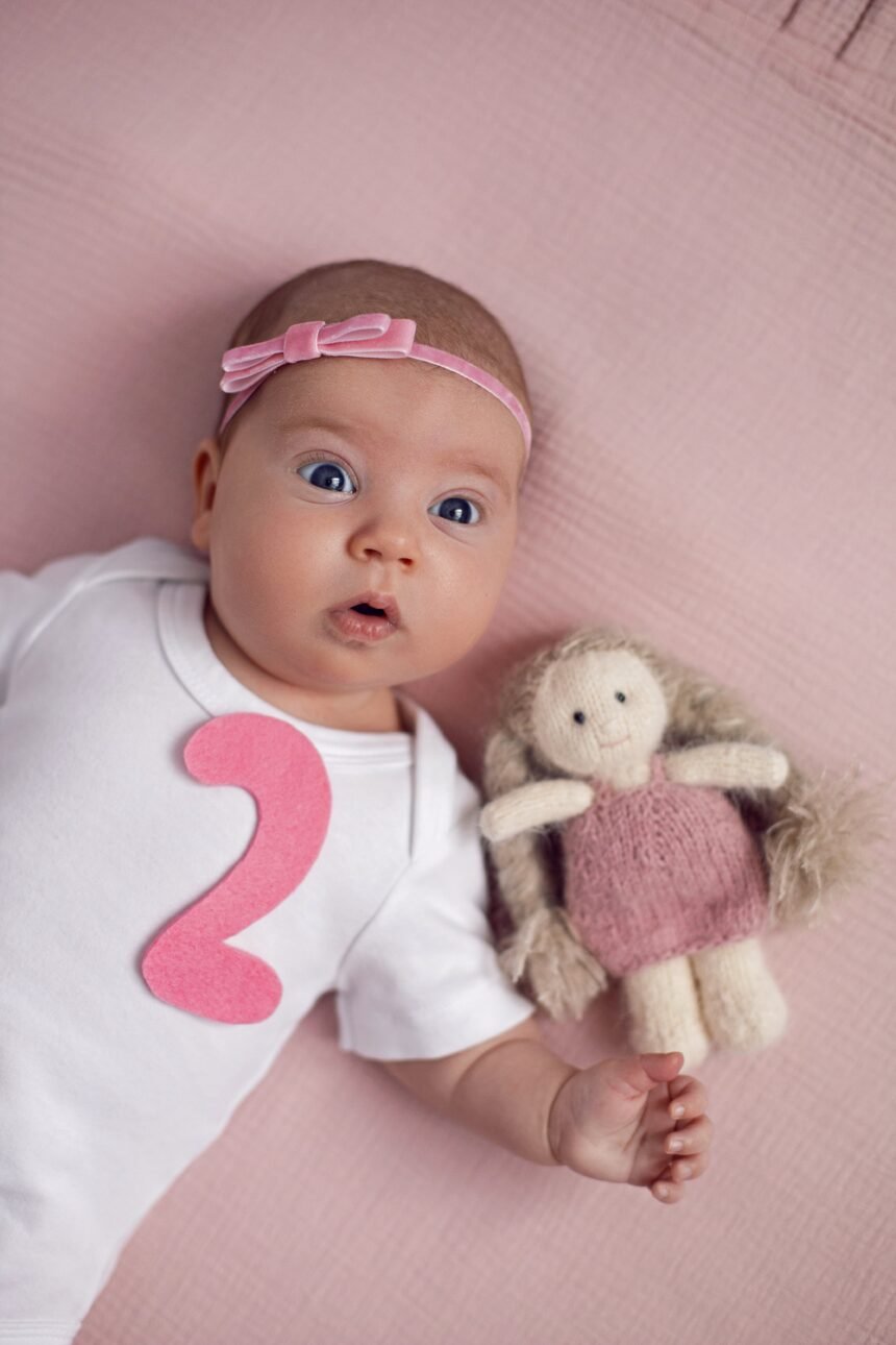 girl baby infant with a bow lies on the bed in a white bodysuit with a doll toy next to it, the child has a pink number 2 months on a pink background.